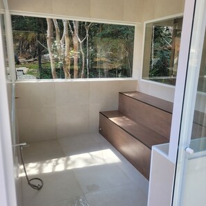 Un baño moderno con cabina de ducha de vidrio, un banco de madera y vista a un bosque a través de un ventanal grande.