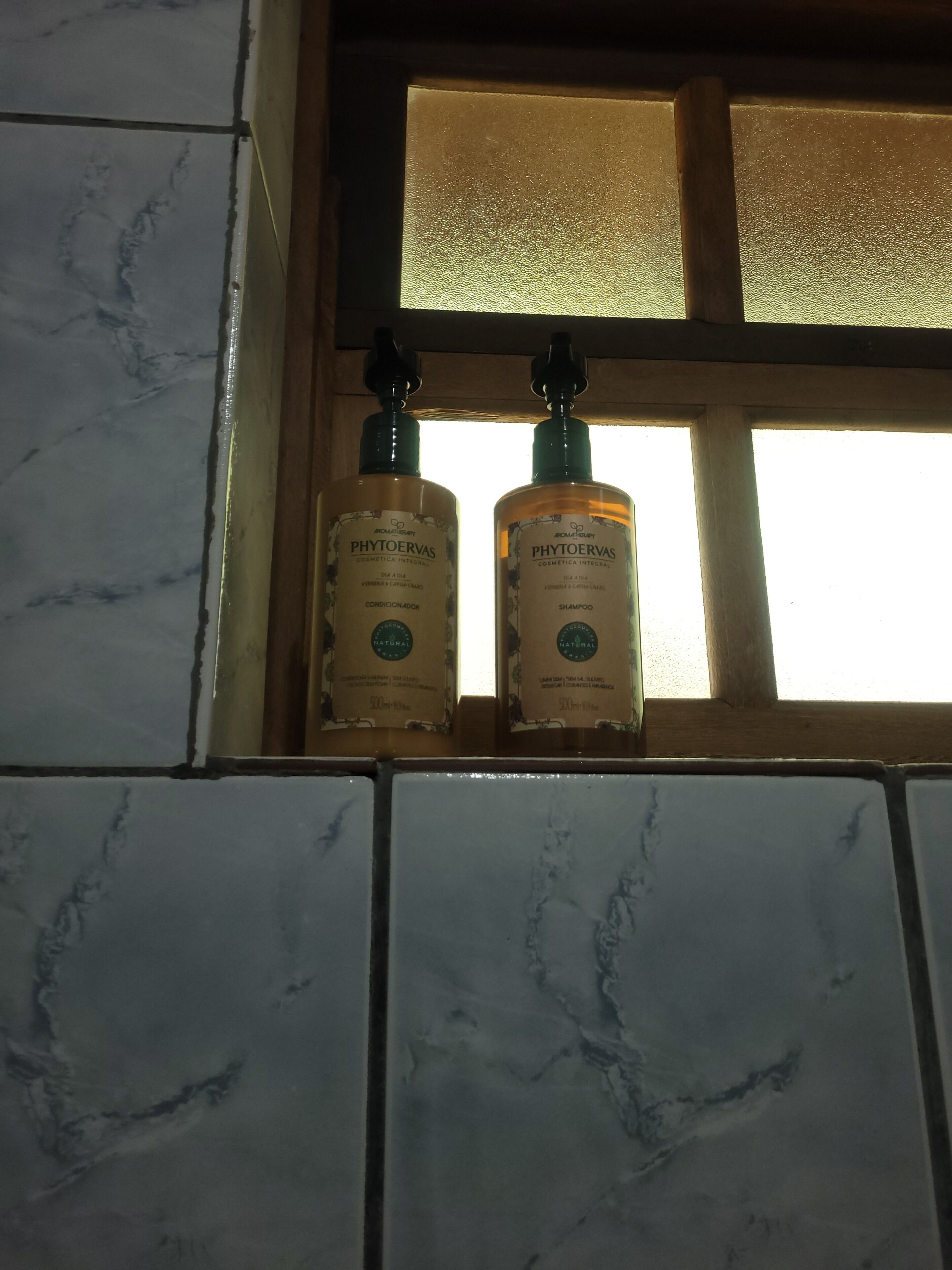 Dos ampolles de xampú Phytoervas en un bany amb rajoles de marbre i una finestra amb vidres motllurats.