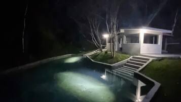 Ein Nachtbild von einem modernen Haus mit Swimmingpool, umgeben von Bäumen und einem gut beleuchteten Weg zum Eingang.
