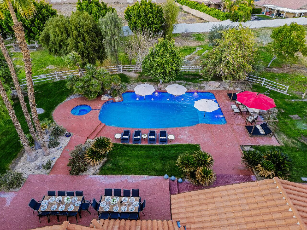 An aerial view of a backyard with a swimming pool, sun loungers, a dining area, and surrounding greenery.