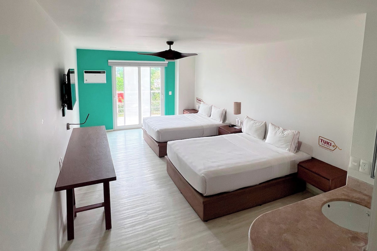 A modern hotel room with two beds, a desk, a small table, a round seating area, and a view of the outdoors.
