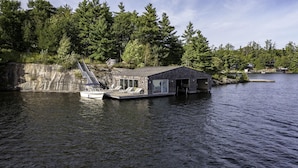 A floating dock with a small building, a boat, and a forested shoreline. - Unique 6 bed 5 bath cottage on Lake Muskoka with Pool (Gravenhurst)