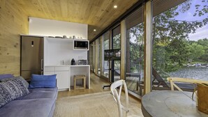 A modern interior with a sofa, dining table, kitchen area, and large windows overlooking a body of water. - Unique 6 bed 5 bath cottage on Lake Muskoka with Pool (Gravenhurst)