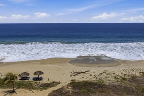 A beach with a clear view of the ocean, a sandy area with a few palm trees, and a small structure with chairs. - One Bedroom Oceanfront - Puerto Escondido 1352 (Bajos de Chila)