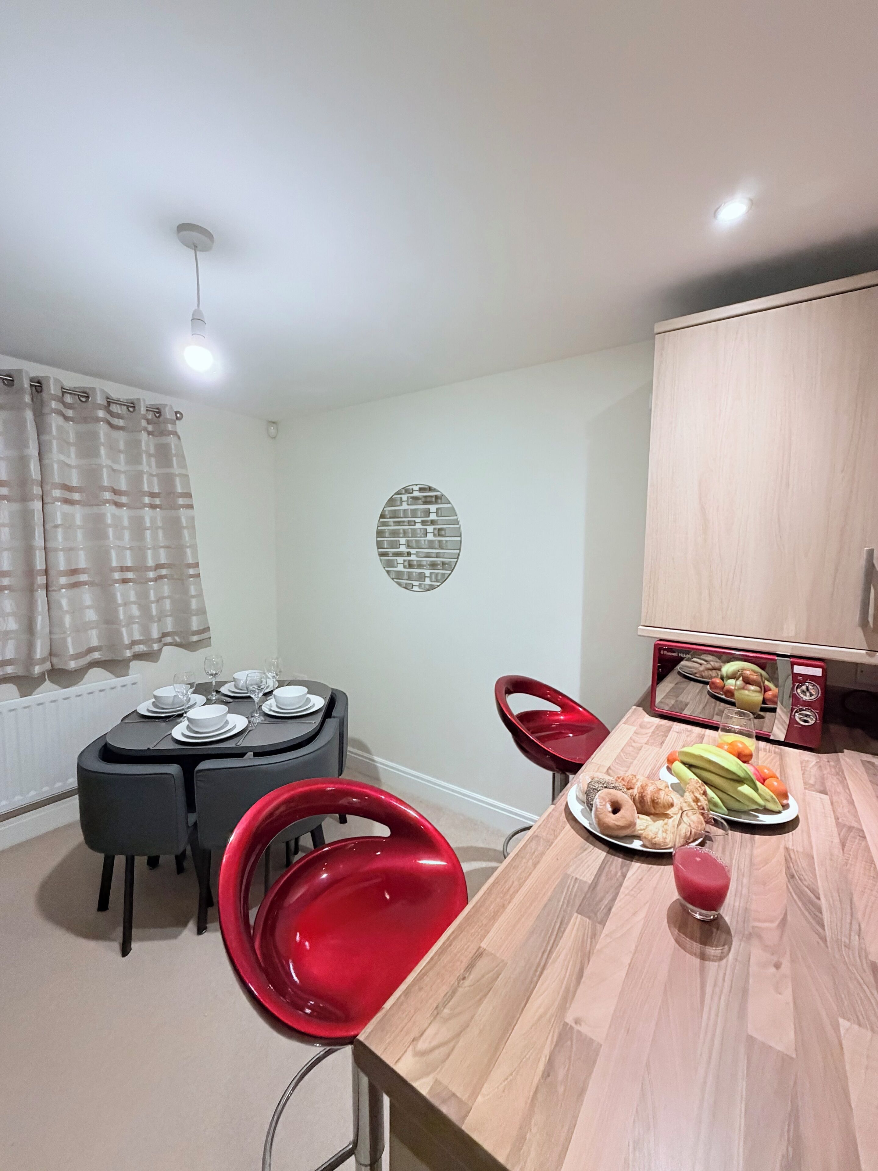 A dining area with a table set for four, a red chair, a microwave, and a fruit plate.
