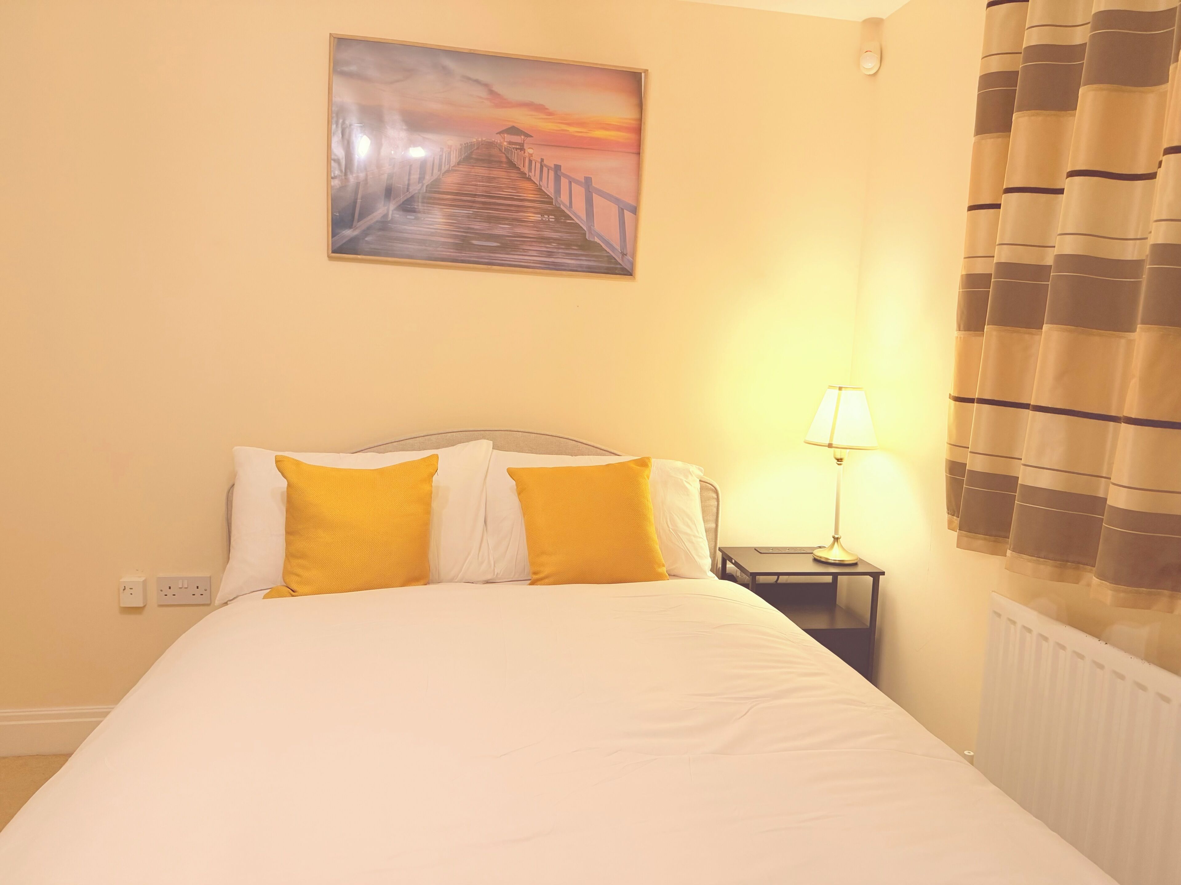 Ein Hotelzimmer mit einem Bett, einem Bild eines Piers, einer Lampe und einem Vorhang.