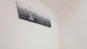 Ein ordentlich bezogenes Bett mit weißen und gelben Kissen, ein schwarzer Nachttisch mit Steckleiste und ein Stadtbild an der Wand.
