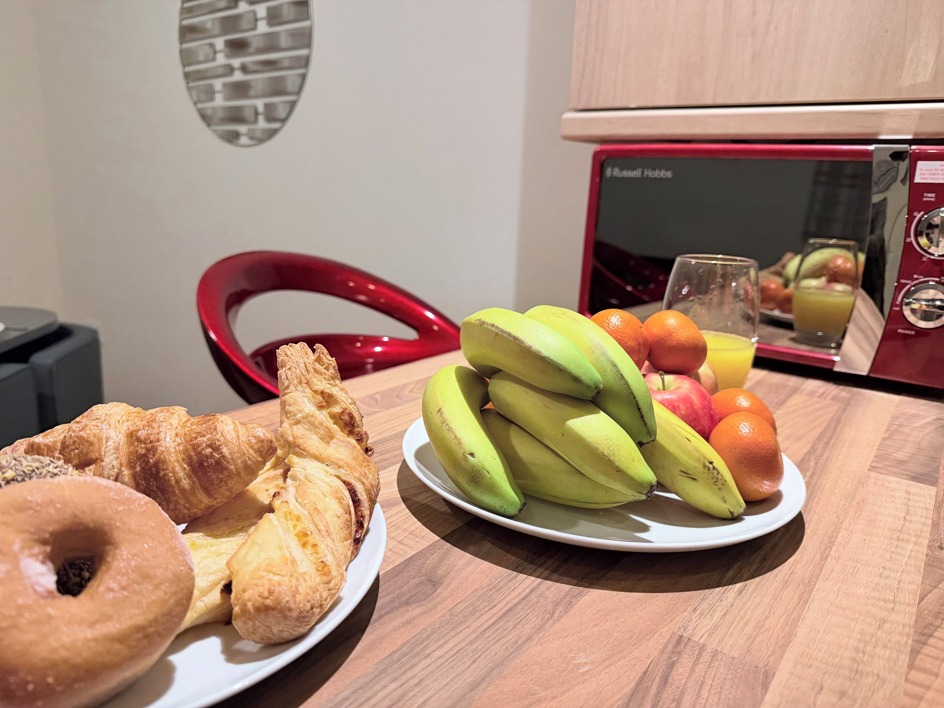 Aamiaispöytä, jossa on croissantteja, banaaneja, omenoita, appelsiineja ja lasi mehua puisella pöydällä.