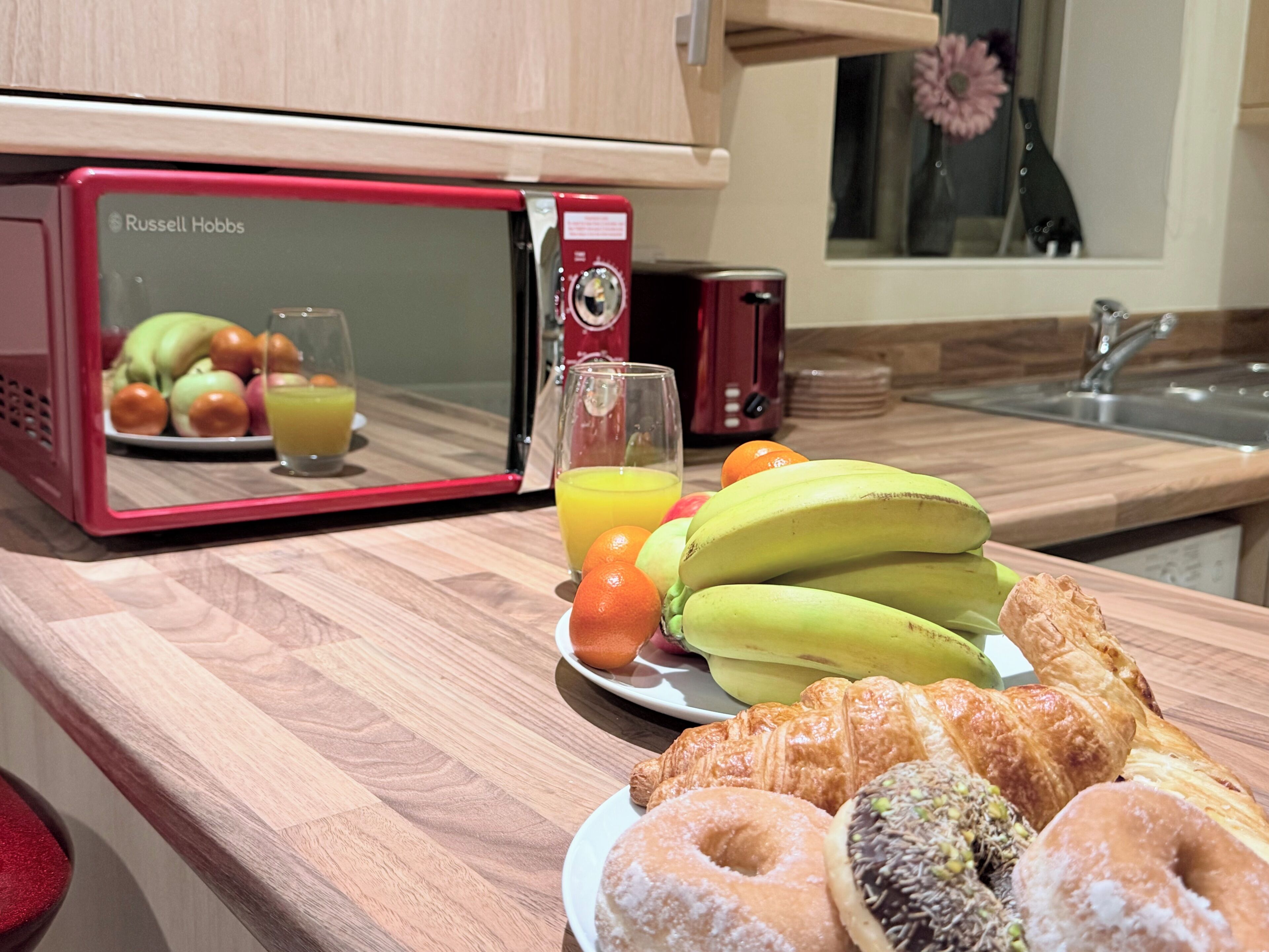 Een aanrecht met een rode magnetron, een bord met bananen, sinaasappels en gebak.