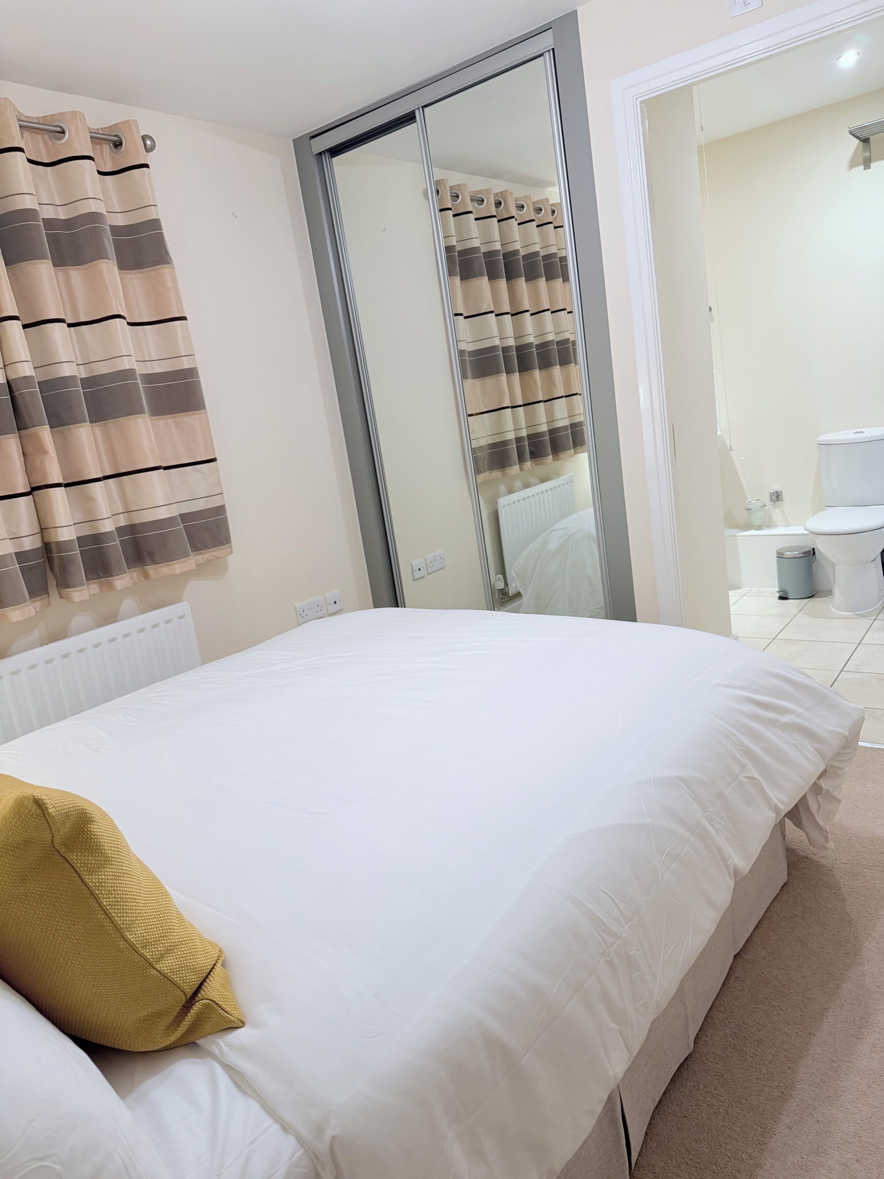 Un dormitorio con cama, espejo, radiador y baño visible a través de una puerta abierta.