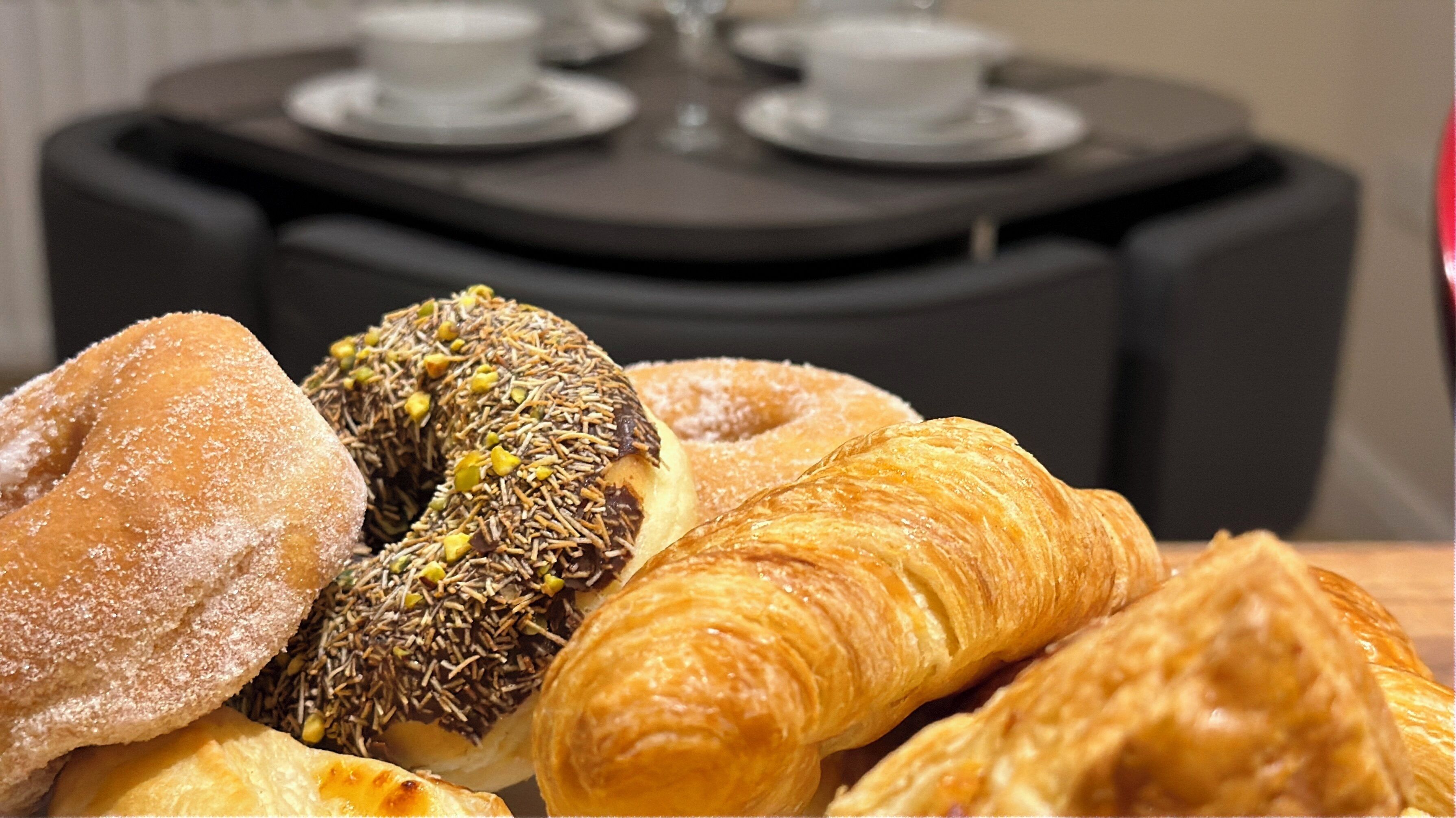 Una mesa con diversos pasteles, entre ellos un donut y croissants, y una vajilla con tazas y platitos.