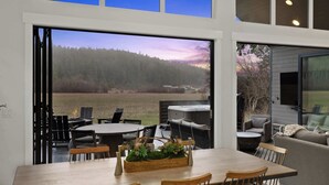 Una zona de menjador amb una taula, cadires i vista a un bosc per grans finestres.