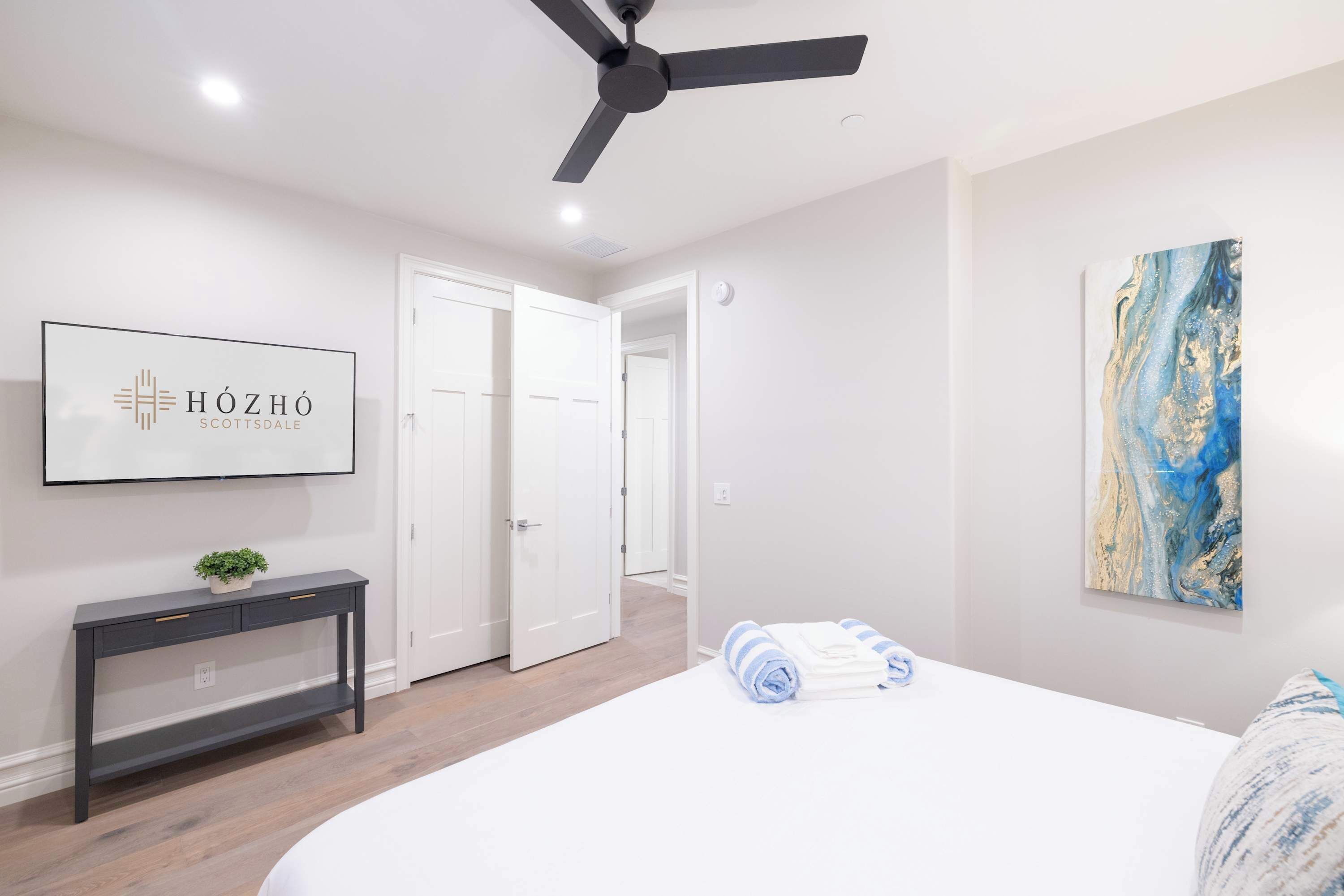 A bedroom with a bed, a ceiling fan, a console table with drawers, a wall-mounted artwork, and a door leading to another room.