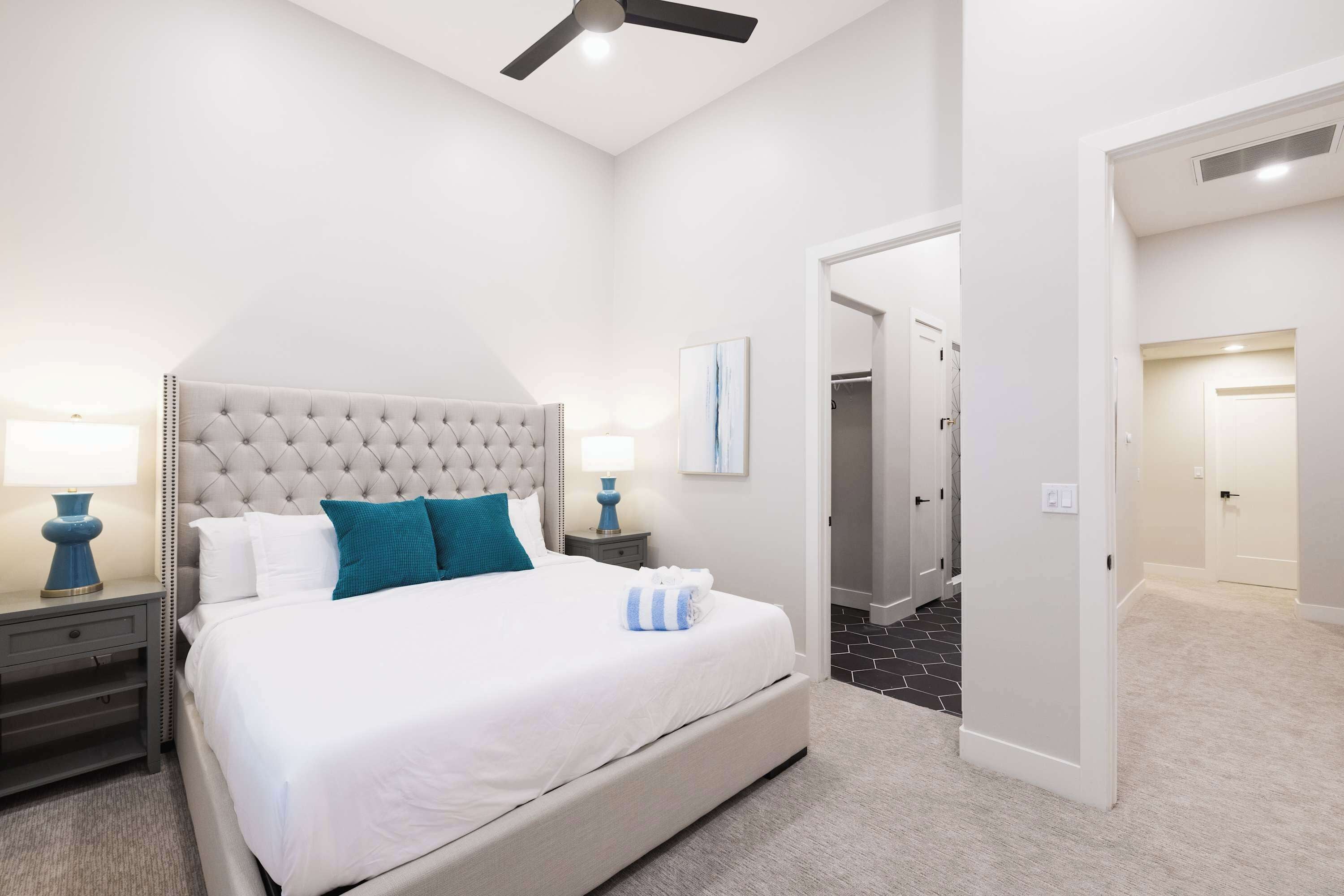 Une chambre à coucher avec une tête de lit capitonnée, un lit avec une literie blanche et turquoise, une table de chevet avec une lampe, et une porte donnant sur une autre pièce.
