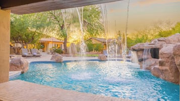 Una piscina con varias fuentes de agua, incluyendo cascadas y una fuente, rodeada de rocas y sillas de descanso.