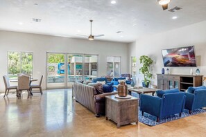 A spacious living room with a large sofa, a coffee table, and a flat-screen TV. There are large windows and a ceiling fan. - HÓZHÓ Cactus | Pool | Pickleball | Theatre | BBQ (Scottsdale)