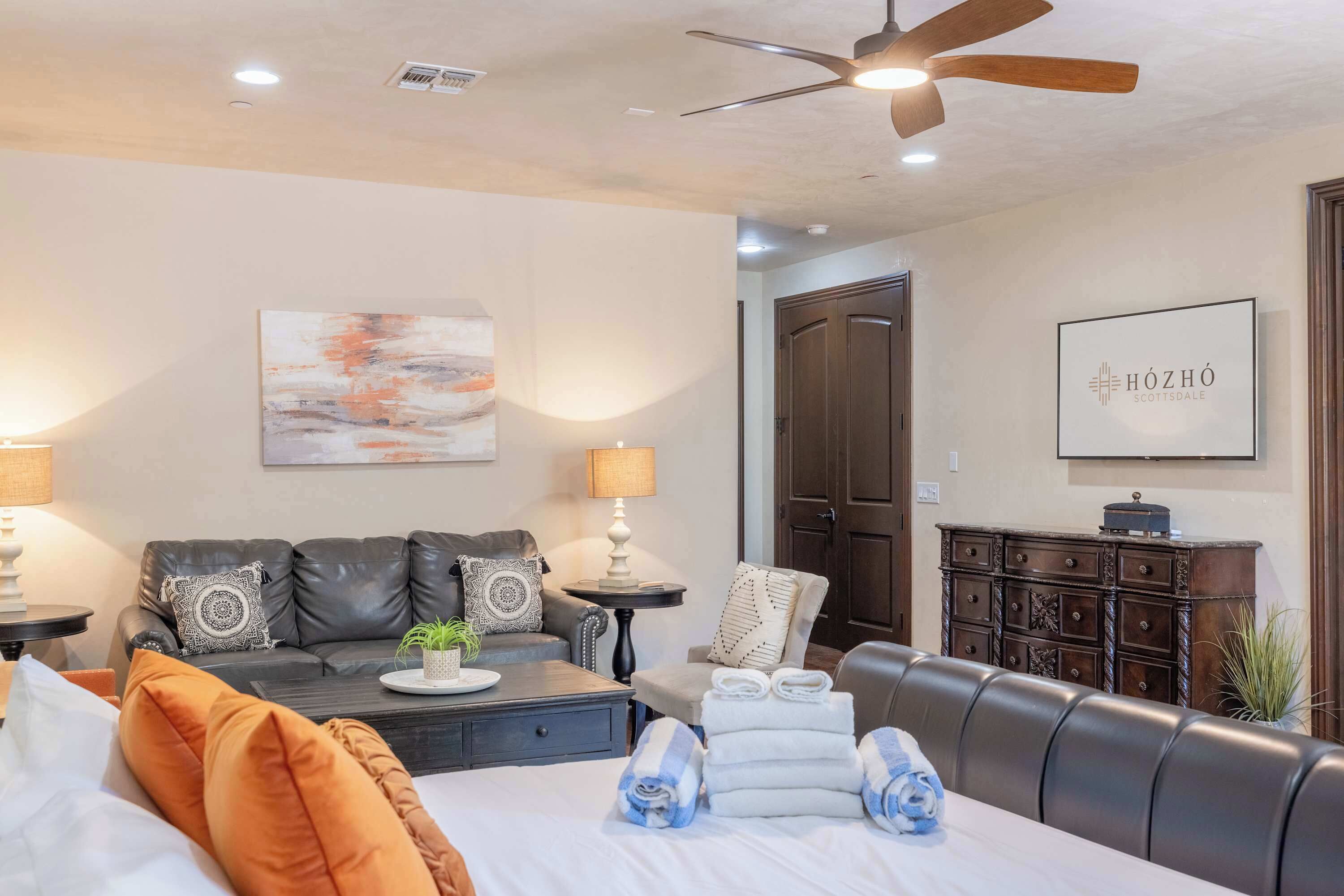 A modern hotel room with a leather sofa, a dining table, a bed with towels, a ceiling fan, and a wall-mounted artwork.