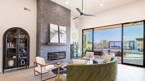 A modern living room with a fireplace, a curved green sofa, a glass cabinet, and a large window offering a view of the outdoors. - HÓZHÓ | Pool, Hot Tub, Theatre, Sauna & Fire Pit (Scottsdale)