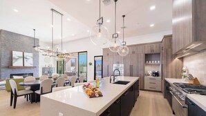 A modern kitchen with a central island, pendant lights, and a dining area with a green sofa and chairs. - HÓZHÓ | Pool, Hot Tub, Theatre, Sauna & Fire Pit (Scottsdale)