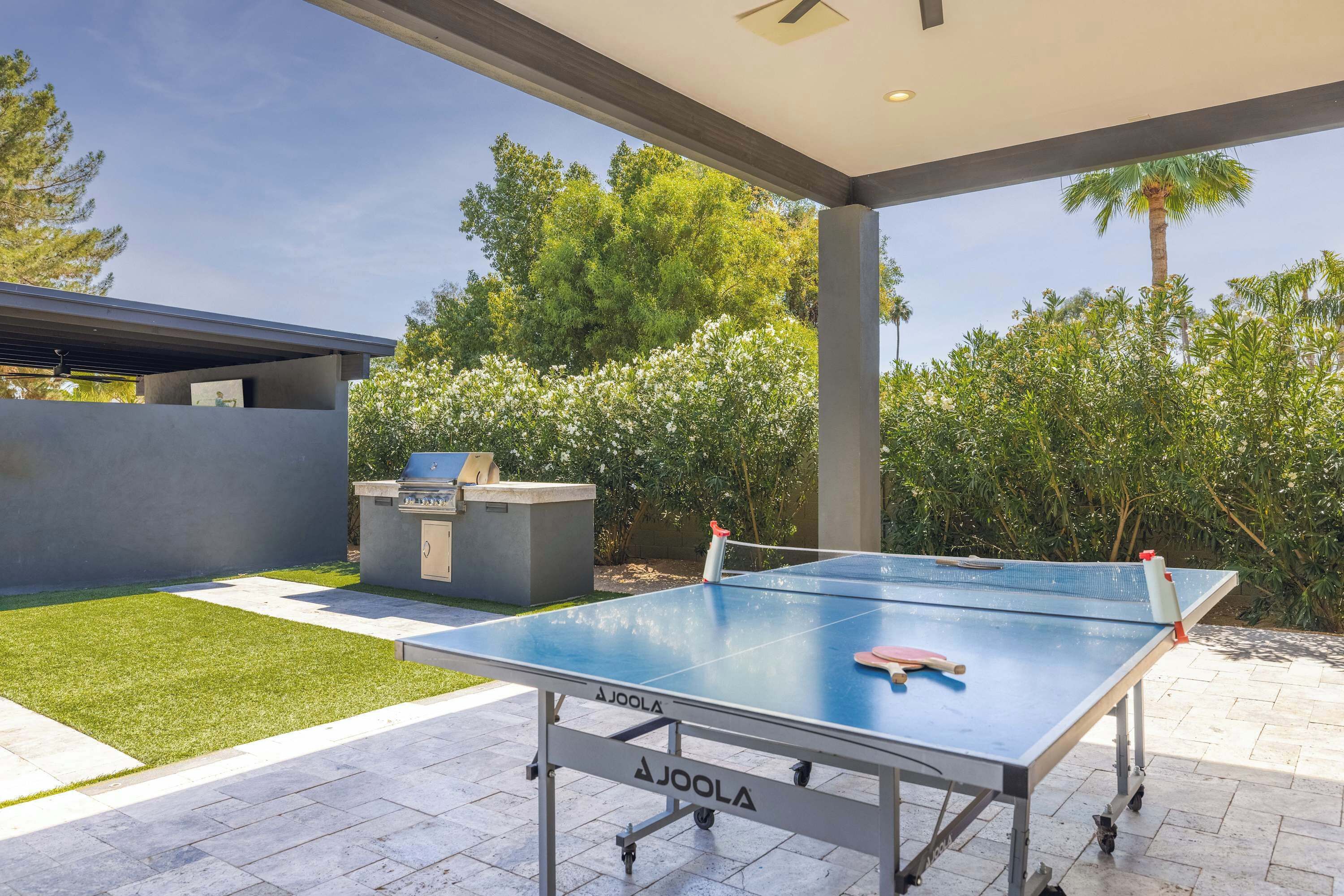 Mesa de ping pong al aire libre con una parrilla y una estructura de concreto en un jardín ajardinado.