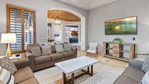 Un salon avec un canapé, une table basse et un téléviseur. On y trouve un meuble en bois orné de plantes et un tableau accroché au mur.