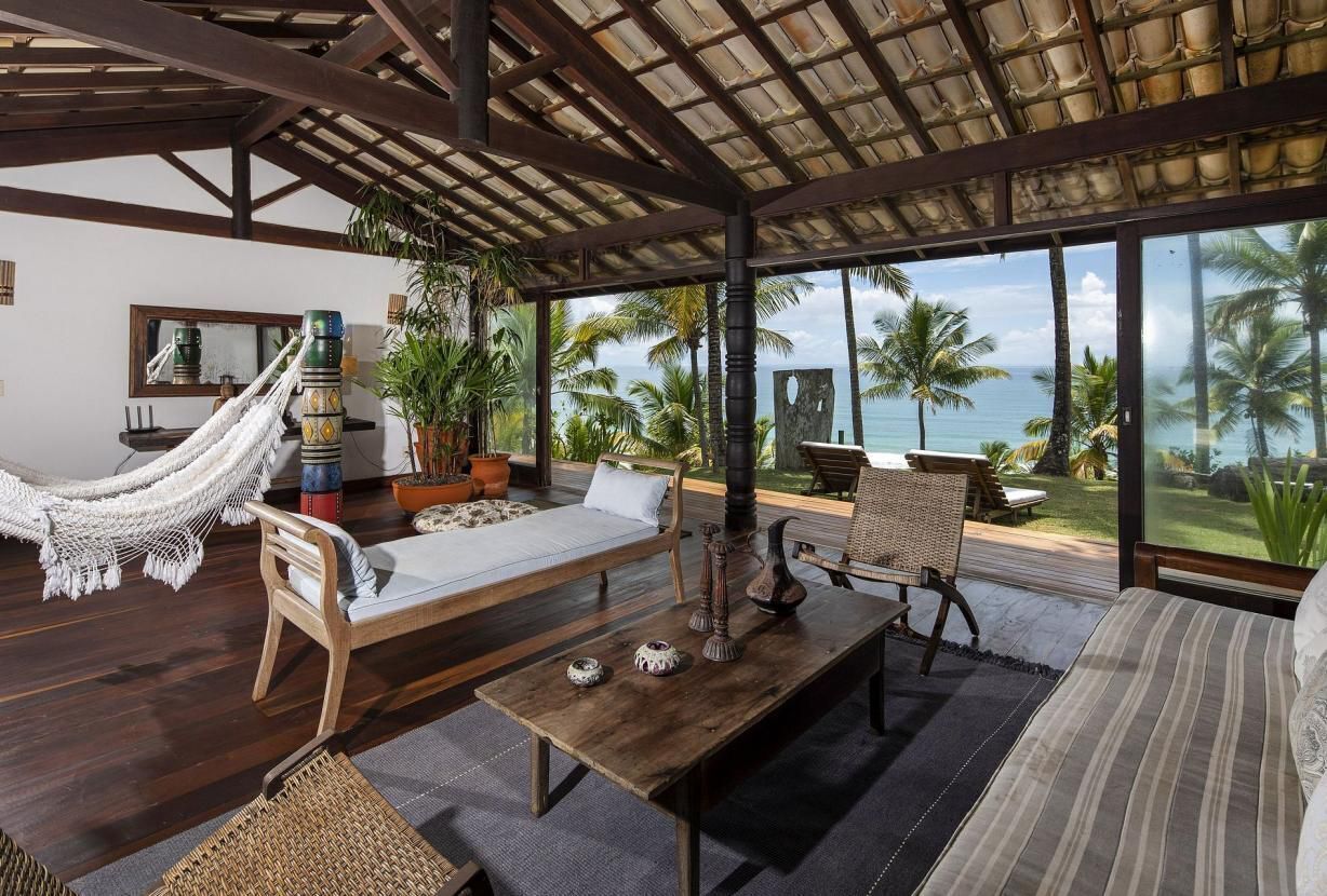 Amplia habitación con hamaca, muebles de madera, vista al océano y plantas tropicales.
