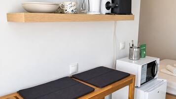 Una kitchenette compacta con microondas, cafetera y un refrigerador pequeño. Hay taburetes de madera y un banco con un tapete oscuro.