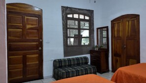 A room with a large window, wooden doors, a bench, a dresser, and two beds with orange bedding.