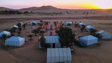 Un resort en el desierto con varias tiendas de techo azul, palmeras y un camino central rojo.