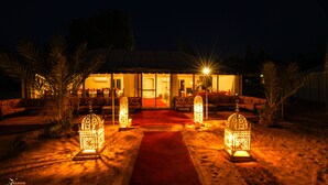 A building with a red carpet leading to a glass door entrance, surrounded by palm trees and illuminated by lanterns. - Merzouga Desert Camp (Merzouga)