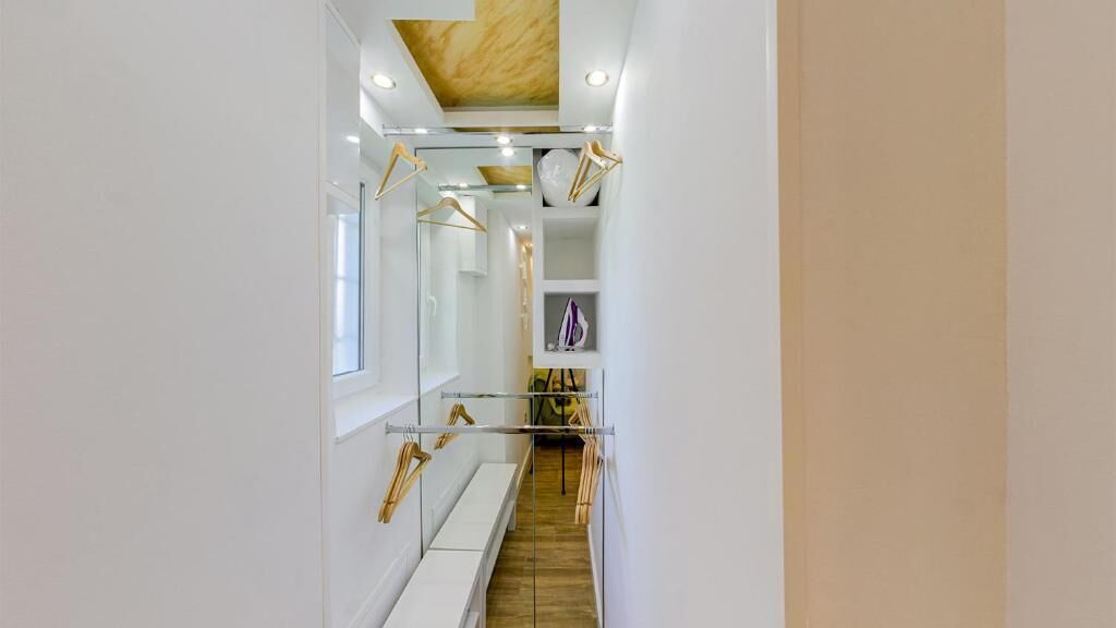 Un couloir étroit avec un banc en bois, un portant à vêtements et une étagère remplie d’objets.