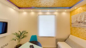 Une pièce avec un plafond orné d’un motif texturé, une fenêtre avec des stores, un téléviseur fixé au mur, une plante, une table avec une télécommande et un canapé.