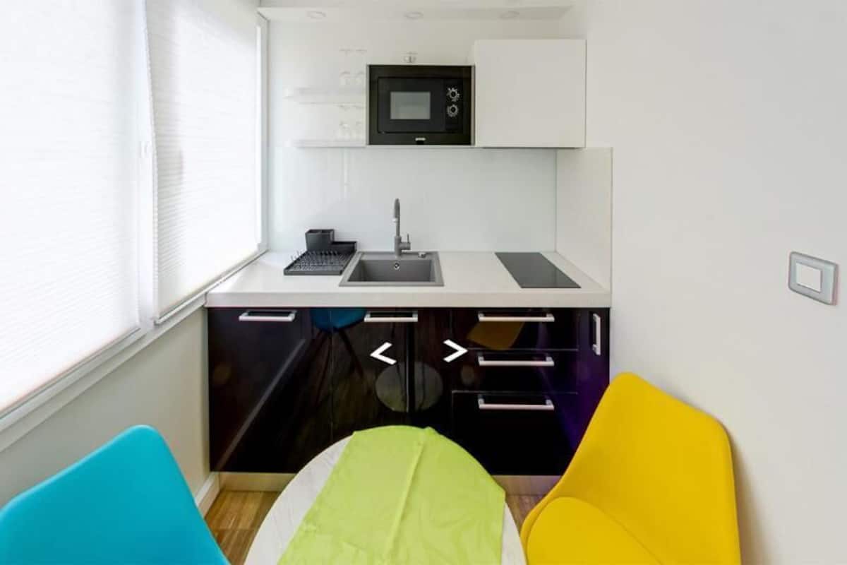 A compact kitchenette with a microwave, sink, and storage drawers, a window with blinds, and two colorful chairs.