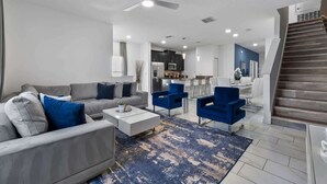 Un soggiorno moderno con divano, tavolino e poltrone blu, una zona cucina con pensili ed elettrodomestici, e una scala.