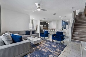Ein modernes Wohnzimmer mit einer Couch, einem Couchtisch und blauen Sesseln, ein Küchenbereich mit Schränken und Geräten sowie eine Treppe.
