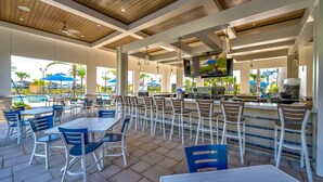 Area pranzo all'aperto con tavoli e sedie, un bar e una piscina sullo sfondo.
