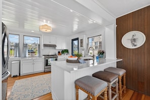 A modern kitchen with white cabinetry, a central island with stools, a stainless steel refrigerator, and a wooden accent wall. - Oceanfront with Views! Sleeps 10, Central AC! (Scituate)