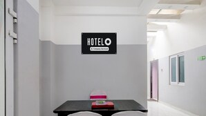 Zona de recepción de hotel con un cartel que indica que es un hotel gestionado por una empresa. Hay dos sillas con lazos rosas, una mesa negra con una carpeta roja y una pared blanca.
