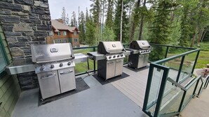 Een balkon met drie barbecuegrills, een stenen muur en uitzicht op een huis en bos.
