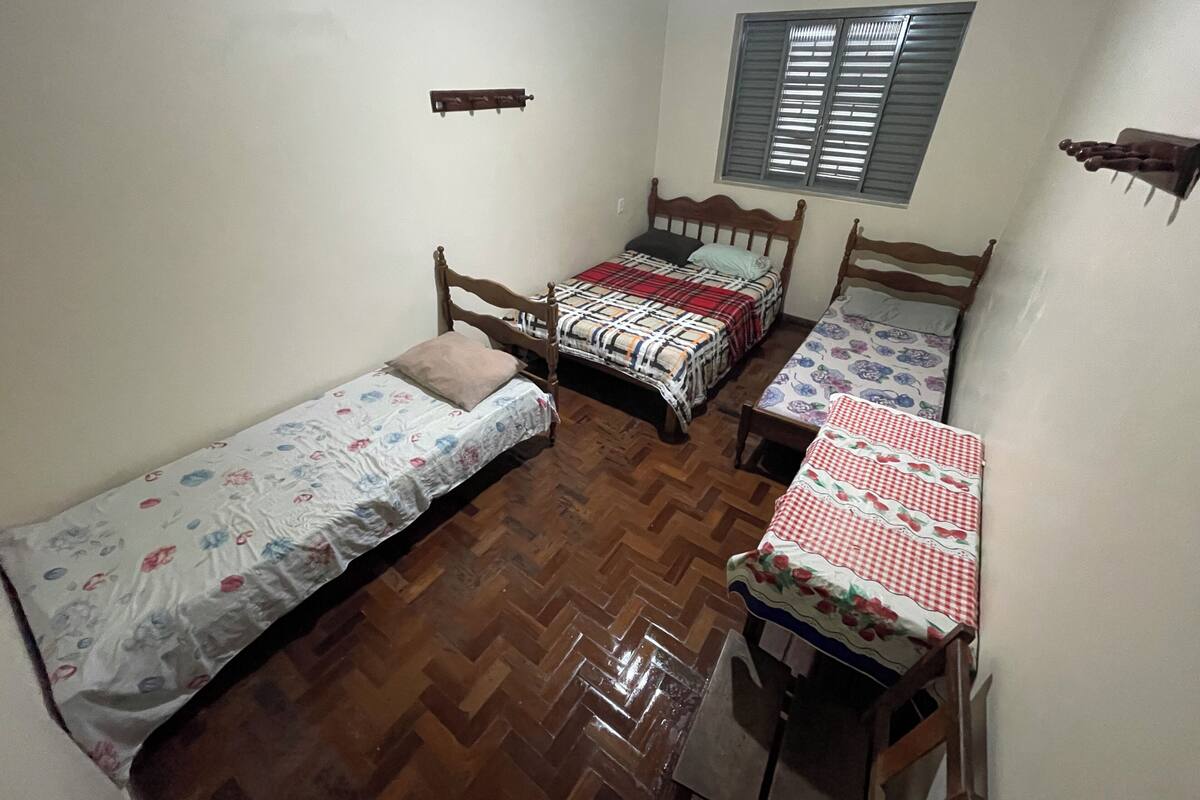 A dormitory room with three wooden beds, a window with blinds, a wooden floor, and a small shelf.