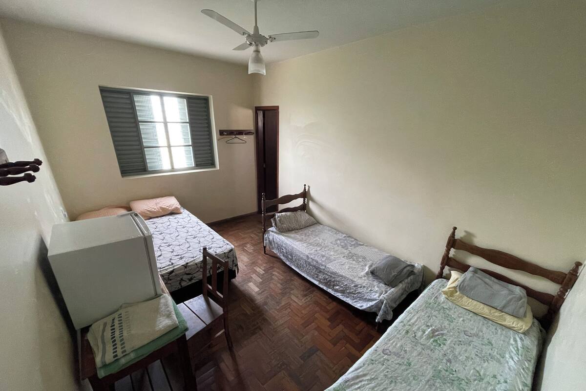 A dormitory room with two single beds, a desk with a chair, a ceiling fan, and a window with blinds.