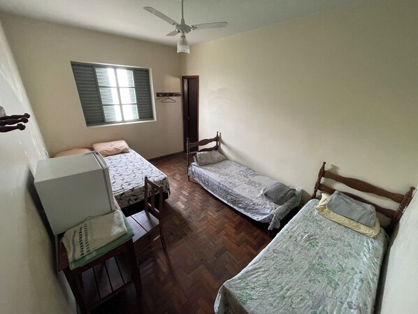 A dormitory room with two single beds, a desk with a chair, a ceiling fan, and a window with blinds. - Hotel São José (Patos de Minas)
