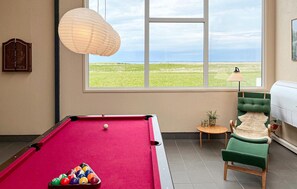 Una mesa de billar con bolas, una ventana que da a un campo, una lámpara y una silla verde.