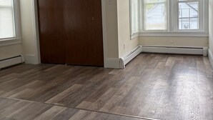 Ein Zimmer mit Holzdielenboden, einer großen, dunklen Holztür und einem Fenster mit Jalousien.