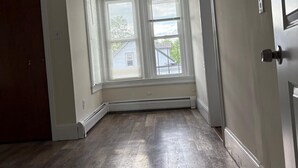 Ein Zimmer mit Holzboden, einem großen Fenster mit Jalousien und einer Tür.