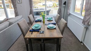 Zona pranzo con tavolo imbandito per quattro persone, divano e vista su un'area parcheggio attraverso ampie finestre.