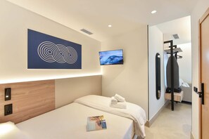 Ein Hotelzimmer mit Bett, Wandfernseher, Handtuch auf dem Bett, Holz-Kopfteil und einer Tür zu einem Badezimmer.