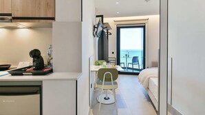 Et aflangt hotelværelse med tekøkken, et spiseområde med bord og stole, en seng og en balkon med havudsigt.