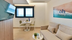 Un salon moderne avec un canapé, une petite table ronde agrémentée d’une plante, un téléviseur fixé au mur et une fenêtre donnant sur la mer.