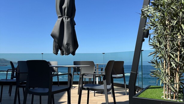 Un espace extérieur aménagé avec des tables et des chaises, un parasol noir et une vue sur l’océan.